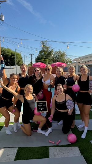 Ten women in black workout outfits posing by a sunny backyard pool on a residential patio, holding pink exercise balls, small dumbbells, a champagne bottle, face cutouts and a letterboard — playful outdoor fitness-style pool party.