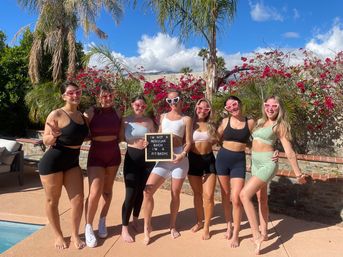Seven women in colorful activewear and heart-shaped sunglasses posing poolside in a sunny backyard with palm trees and pink bougainvillea; center woman holds sign reading "I'M NOT A REGULAR BACH I'M A FIT BACH!"