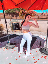 Smiling woman in white leggings flexes a bicep under a red patio umbrella on a sunny backyard, holding a board reading "I'M NOT A REGULAR BACH I'M A FIT BACH" with yoga mats and rose petals scattered around.