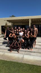 Outdoor yoga group photo: people in activewear and one in white posing barefoot on sunlit patio steps under a pergola by a stucco building and palm tree — fitness retreat vibe.
