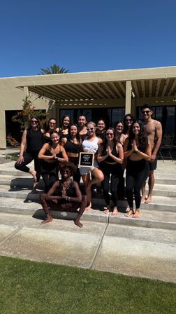 Outdoor yoga group photo: people in activewear and one in white posing barefoot on sunlit patio steps under a pergola by a stucco building and palm tree — fitness retreat vibe.