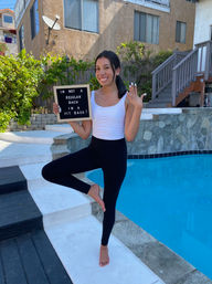 Smiling woman in a white tank and black leggings balances on one leg by a backyard pool, holding a letter board that reads "I'M NOT A REGULAR BACH I'M A FIT BACH!"