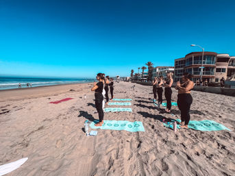Group beach yoga on turquoise mats along a sunny oceanfront with palm trees and beachfront homes under a clear blue sky
