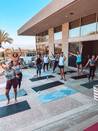 Group of women in a sunny outdoor yoga class on a poolside patio with mats, palm trees and distant mountains, smiling and waving