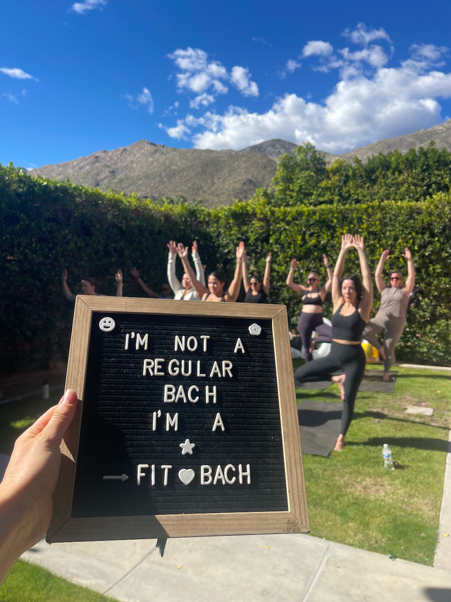 Hand-held letter board reading 'I'm not a regular bach I'm a fit bach' in the foreground; group of women practicing outdoor yoga on a sunny lawn with a hedge and mountain backdrop.