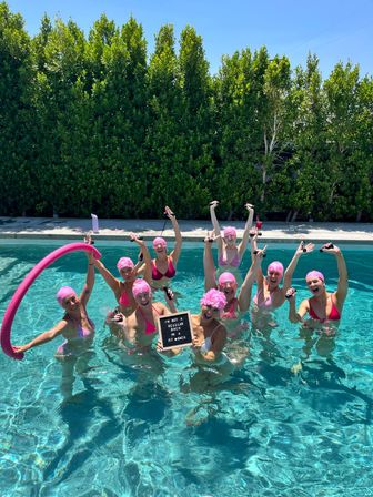 Group of women in pink swim caps and bikinis doing a fun aqua workout in a sunny backyard pool, cheering with a pink pool noodle, handheld water weights and a small letter board.