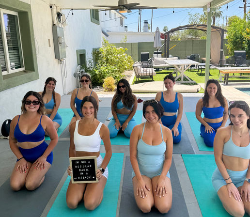 Group of women in blue activewear kneeling on yoga mats under a covered backyard patio beside a pool, smiling for a group fitness photo; one holds a sign reading "I'm not a regular bach, I'm a fitbach!"