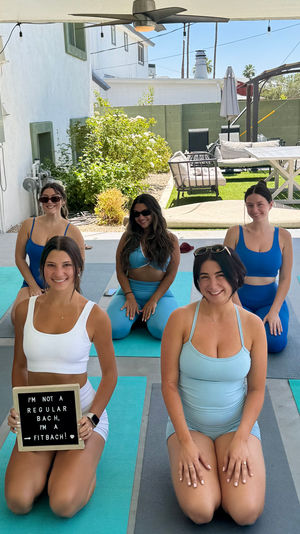Group of women in blue activewear kneeling on yoga mats under a covered backyard patio beside a pool, smiling for a group fitness photo; one holds a sign reading "I'm not a regular bach, I'm a fitbach!"
