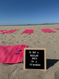 Sunny sandy beach set up for a beach workout — rows of pink yoga mats and resistance bands, framed letter board in foreground reading "I'm not a regular bach I'm a fit bach", calm ocean and clear blue sky on the horizon.