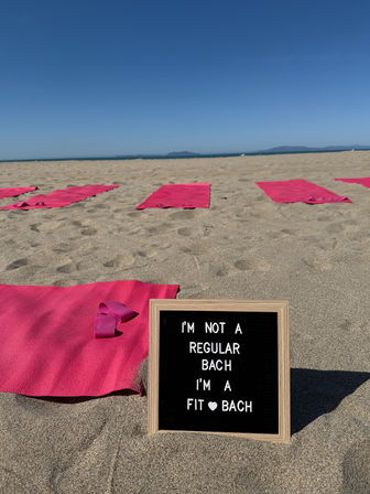 Sunny sandy beach set up for a beach workout — rows of pink yoga mats and resistance bands, framed letter board in foreground reading "I'm not a regular bach I'm a fit bach", calm ocean and clear blue sky on the horizon.
