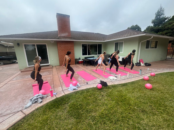 Outdoor pink-themed fitness class on a suburban backyard patio — women on pink yoga mats doing lunges in front of a single-story green house, with pink exercise balls, water bottles and cloudy sky overhead.