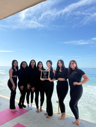 Seven women in black activewear posing barefoot on an oceanfront balcony by the beach, holding a letterboard that reads 'I'M NOT A REGULAR BACH I'M A FIT BACH' with pink yoga mats and a bright blue sky overhead.
