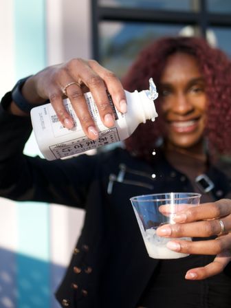 Sunlit close-up of a smiling woman with curly hair pouring a white bottled drink into a plastic cup of ice on an outdoor patio.