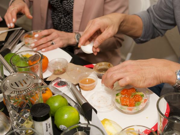 Hands assembling a small poke-style bowl of diced salmon, avocado and sesame on a cluttered dining table with limes, persimmons, sauces and utensils.