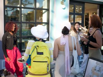 Group of friends chatting outside a sunny urban cafe patio, bright outfits and a yellow backpack, iced drinks and storefront windows