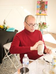 Person in a red turtleneck and glasses shaking a stainless cocktail shaker at a sunny indoor art studio table with plates, a water bottle, and a colorful easel painting in the background.