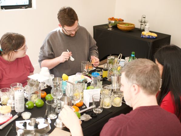 Adults in a hands-on cocktail class preparing drinks around a cluttered table with citrus fruits, glassware, shakers, bitters, and garnishes.