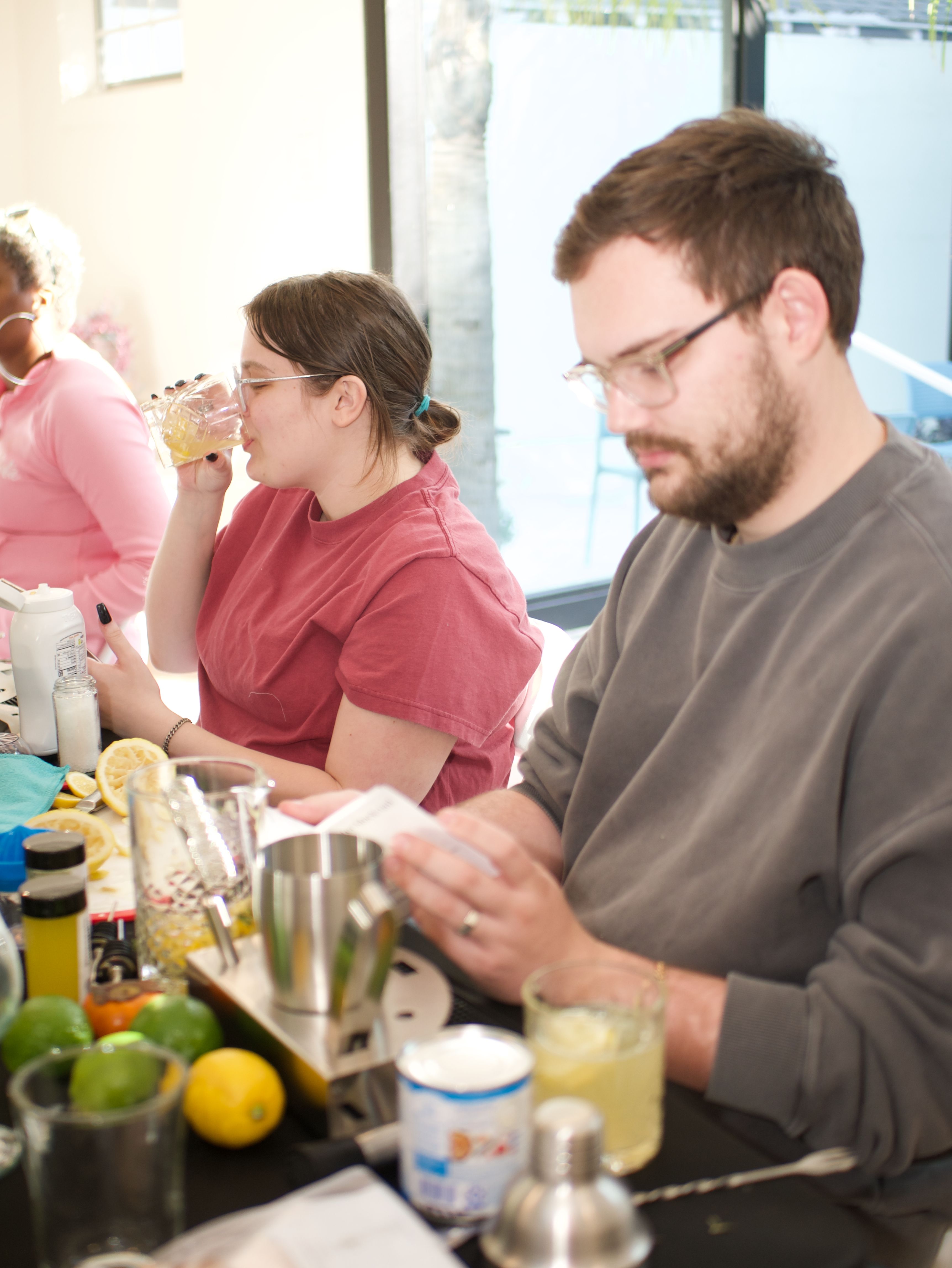 Two people in a sunlit kitchen sampling cocktails at a DIY mixology table with lemons, limes, cocktail shaker and recipe notes