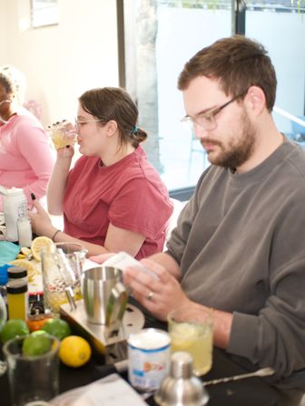Two people in a sunlit kitchen sampling cocktails at a DIY mixology table with lemons, limes, cocktail shaker and recipe notes