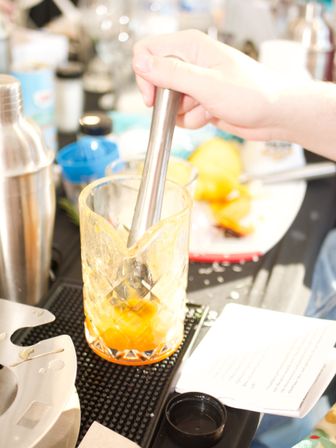 Hand using a stainless-steel muddler to crush orange pieces in a patterned mixing glass on a cocktail bar mat with citrus peels and bartending tools nearby.