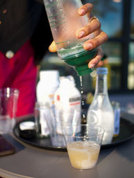 Close-up of a hand squeezing a green-topped bottle as droplets fall into a clear plastic cup of fizzy pale drink, with blurred bottles and cups on a tray in the background.