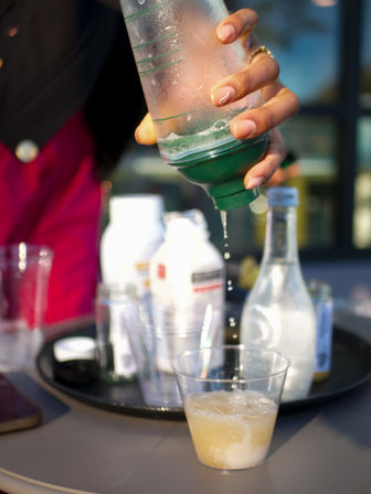 Close-up of a hand squeezing a green-topped bottle as droplets fall into a clear plastic cup of fizzy pale drink, with blurred bottles and cups on a tray in the background.