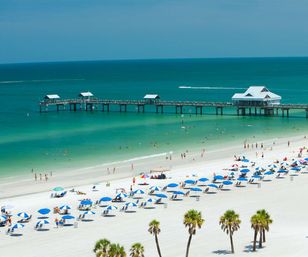 Aerial view of a Gulf Coast white-sand beach with turquoise water, a long wooden pier, rows of blue umbrellas, palm trees and sunbathers.