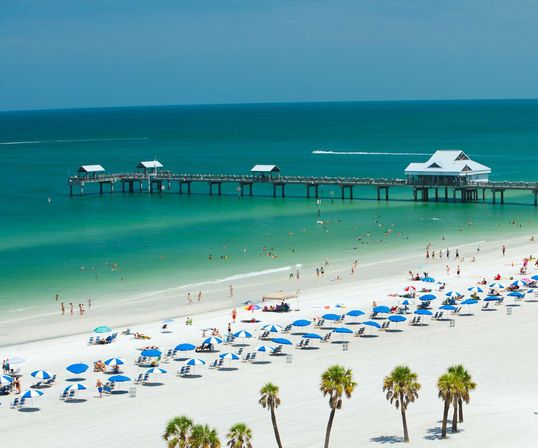 Aerial view of a Gulf Coast white-sand beach with turquoise water, a long wooden pier, rows of blue umbrellas, palm trees and sunbathers.