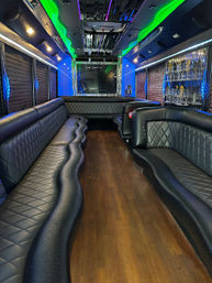 Luxury party bus interior with black quilted leather wraparound seating, wood floor, mirrored ceiling, blue and green LED accent lighting, glassware bar and flat-screen TV.