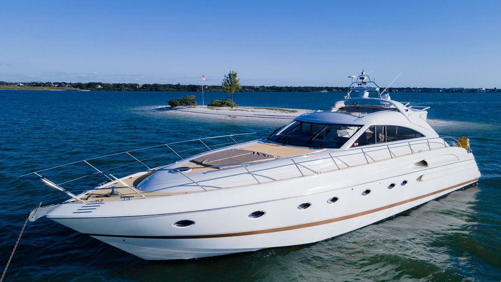 Sleek white luxury motor yacht cruising in clear blue coastal waters past a small sandy island with a flag and trees under a sunny sky.