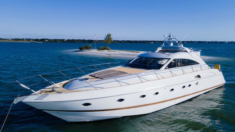 Sleek white luxury motor yacht cruising in clear blue coastal waters past a small sandy island with a flag and trees under a sunny sky.