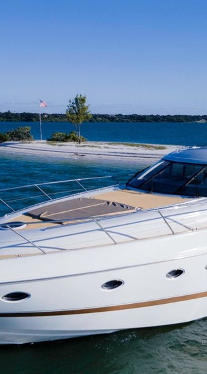 Sleek white luxury motor yacht cruising in clear blue coastal waters past a small sandy island with a flag and trees under a sunny sky.