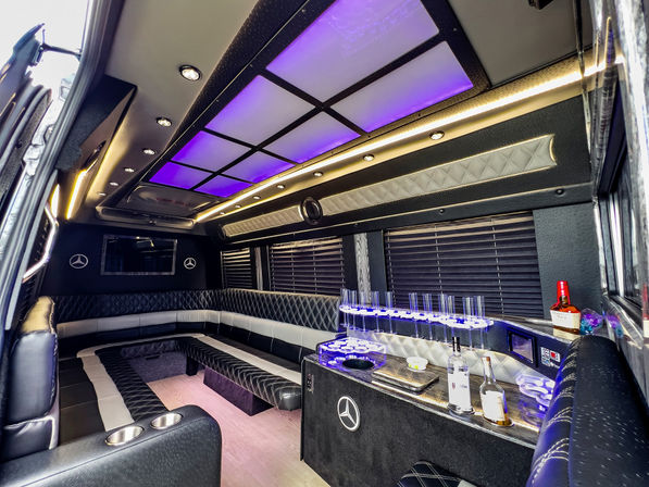 Luxurious black-and-white party limo interior with U-shaped quilted leather seating, purple LED ceiling panels, built-in bar with bottles and glassware, and ambient accent lighting.