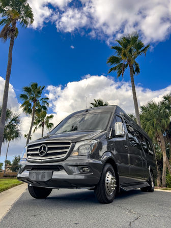 Low-angle shot of a sleek charcoal Mercedes Sprinter van parked on a palm-lined road beneath a bright blue sky and fluffy clouds.
