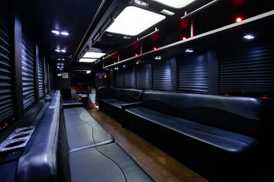 Spacious luxury party bus interior with long black leather bench seats, wood flooring, overhead LED panels and ambient red and white lighting, ready for nightlife group transportation.
