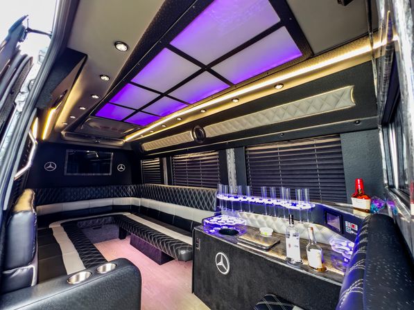 Luxurious party bus interior with purple LED ceiling panels, quilted black-and-white leather wraparound seating, built-in bar with bottles and stemware holders, cup holders and ambient lighting — stylish limo-style shuttle for nightlife and events.