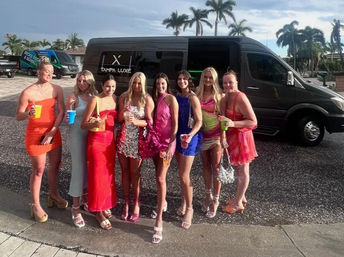 Eight women in colorful cocktail dresses holding drinks pose in front of a black party van on a palm-lined coastal street — girls' night out, party-bus vibe on a sunny afternoon.