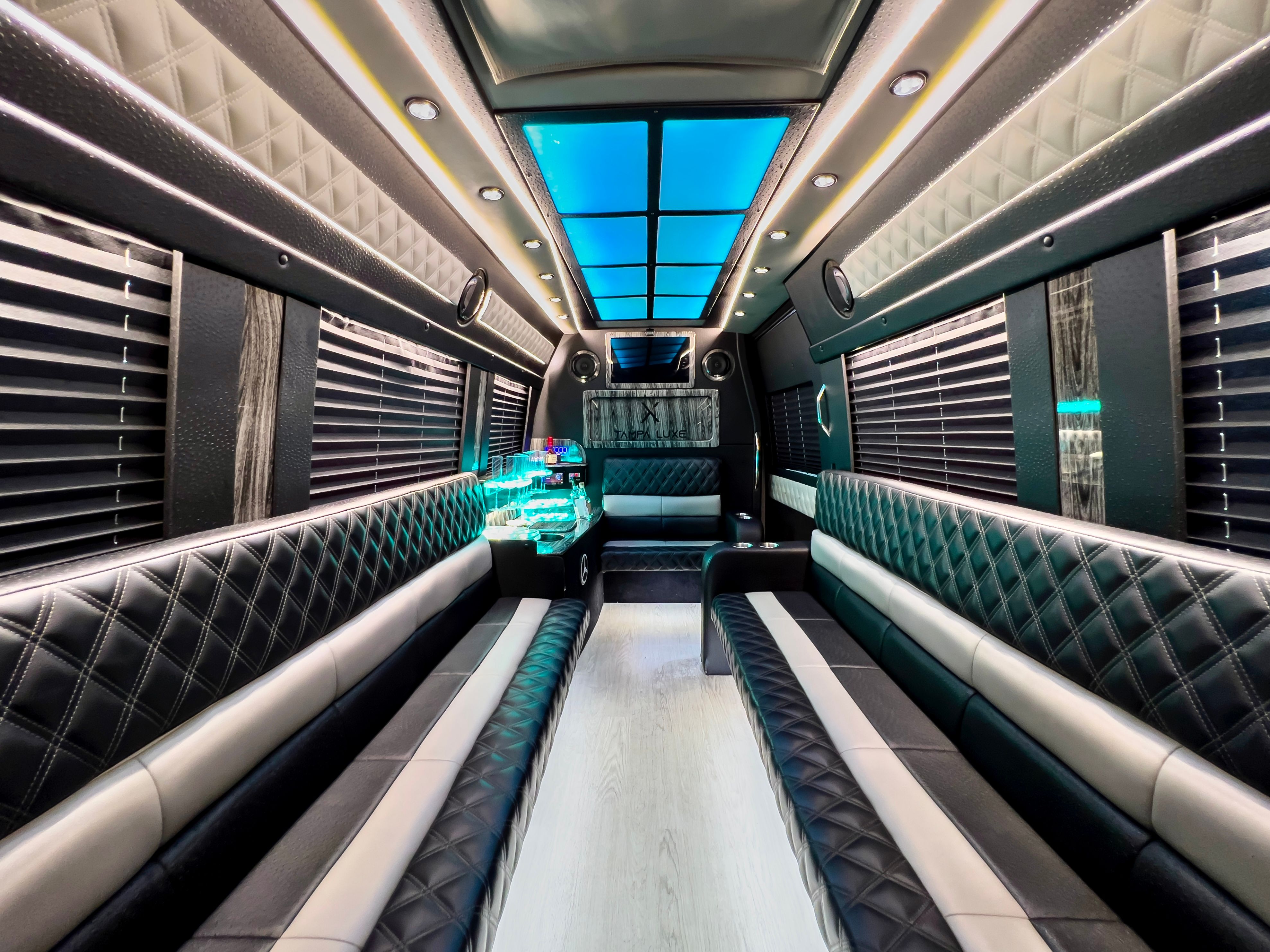 Spacious luxury party bus interior with diamond-stitched black leather bench seats, blue LED ceiling panels, ambient strip lighting, window blinds, and an illuminated mini bar — modern mobile party shuttle.