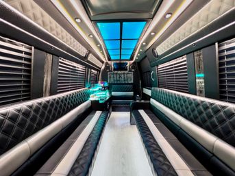Spacious luxury party bus interior with diamond-stitched black leather bench seats, blue LED ceiling panels, ambient strip lighting, window blinds, and an illuminated mini bar — modern mobile party shuttle.
