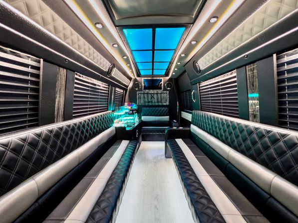 Spacious luxury party bus interior with diamond-stitched black leather bench seats, blue LED ceiling panels, ambient strip lighting, window blinds, and an illuminated mini bar — modern mobile party shuttle.