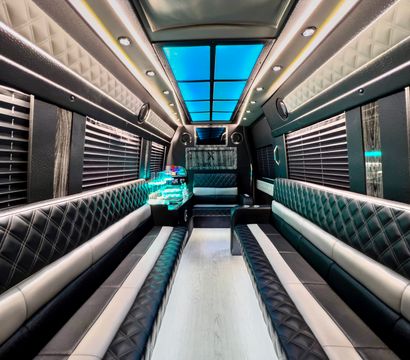 Spacious luxury party bus interior with diamond-stitched black leather bench seats, blue LED ceiling panels, ambient strip lighting, window blinds, and an illuminated mini bar — modern mobile party shuttle.