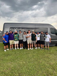 Group of friends posing arm-in-arm in front of a gray passenger van on a grassy field under dark, stormy clouds