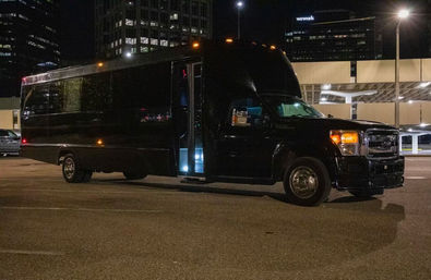 Sleek black shuttle bus parked in a downtown parking lot at night, open side door glowing with blue step light and city high-rises and streetlights in the background