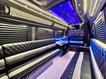 Luxury party bus interior with blue LED ceiling panels, quilted black-and-white leather bench seating, blackout blinds and an illuminated mini bar for VIP event transport.