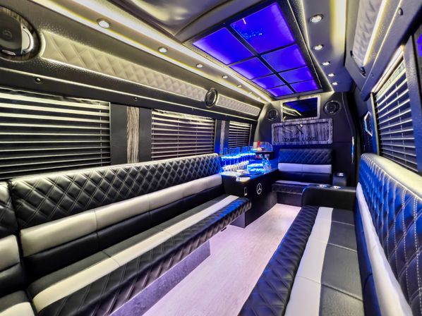 Luxury party bus interior with blue LED ceiling panels, quilted black-and-white leather bench seating, blackout blinds and an illuminated mini bar for VIP event transport.