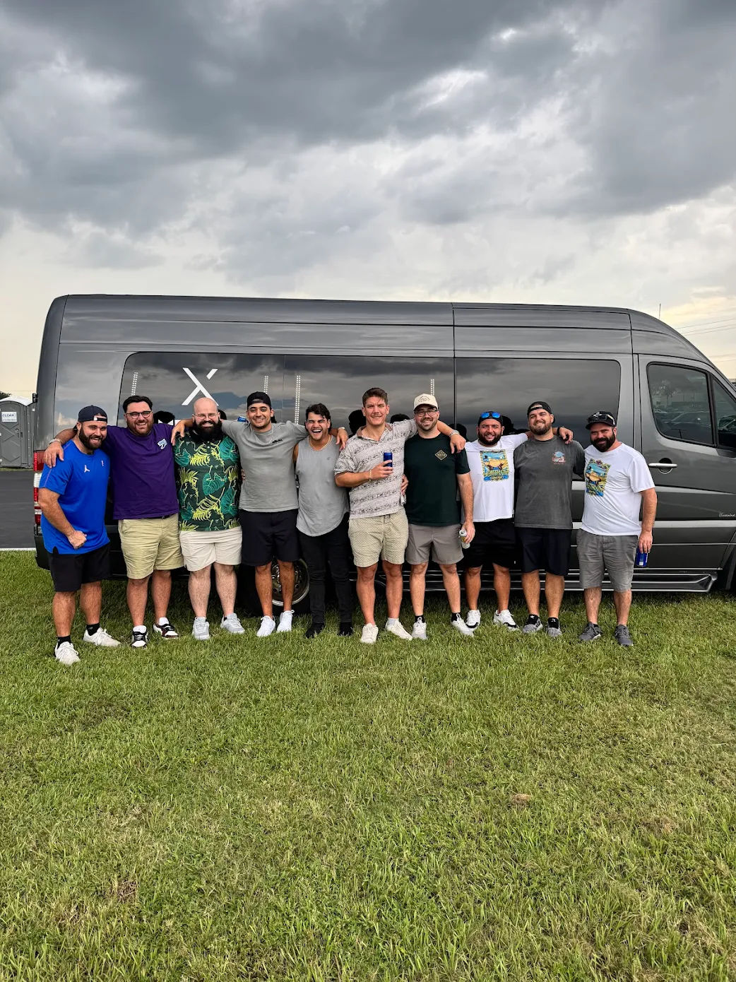 Group of eleven men posing arm-in-arm in front of a gray passenger van on a grassy field under stormy clouds at an outdoor event