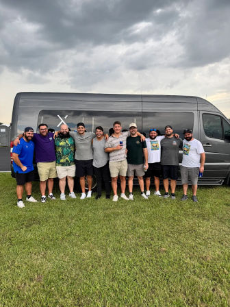 Group of eleven men posing arm-in-arm in front of a gray passenger van on a grassy field under stormy clouds at an outdoor event