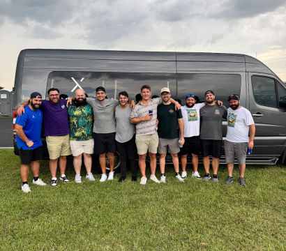 Group of eleven men posing arm-in-arm in front of a gray passenger van on a grassy field under stormy clouds at an outdoor event