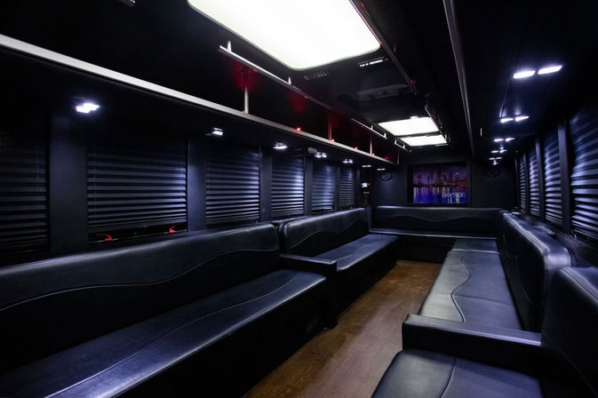Luxury party bus interior with wraparound black leather seating, ambient LED ceiling lights, closed window shades and wood flooring — stylish VIP shuttle for city nightlife.