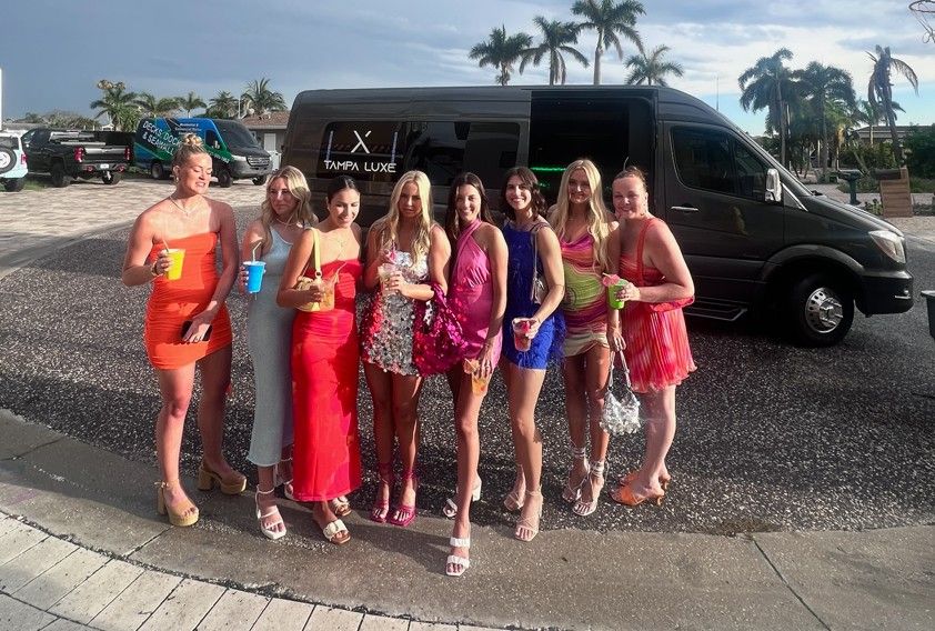 Eight women in colorful cocktail dresses holding drinks and posing in front of a black party shuttle with palm trees and a sunny coastal Florida backdrop.